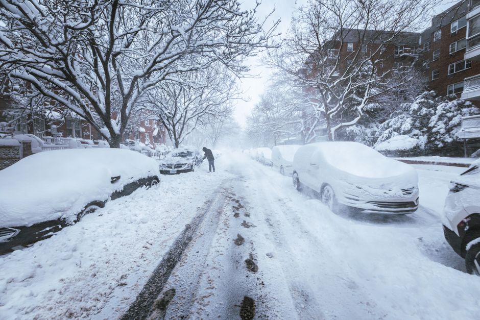 A snowy urban street scene with multiple parked cars covered in a thick layer of snow, including a dark-colored vehicle on the left and a light-colored vehicle on the right, both with snow drifts on their roofs and windshields. The street itself has tire tracks and footprints visible in the snow, indicating recent activity. In the middle distance, a person dressed in dark winter clothing is clearing snow from the road with a snow shovel. Tall, leafless trees with snow-laden branches line both sides of the street, creating a wintery canopy. Residential buildings with brick facades and balconies are visible along the left and right edges, partially obscured by snow and mist. The scene has a diffused, cool-toned lighting typical of overcast winter weather, giving an overall calm and quiet atmosphere. This image emphasizes the importance of effective waste and debris management in snowy conditions, which companies like Waste Removal Queen's Park facilitate through private rubbish removal services when residential streets are affected by severe weather, ensuring safe and accessible streets for disposal needs.