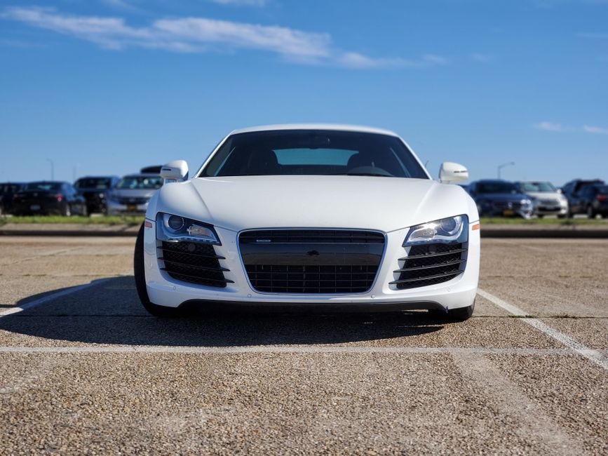 A white sports car with a sleek, aerodynamic design parked in an outdoor lot on a textured asphalt surface, facing directly towards the camera. The vehicle features a large, prominent front grille with black mesh, sharp angular headlights, and smooth, curved body panels with a glossy finish. The car's side mirrors are also white, matching the body color. In the background, a row of other parked vehicles, including sedans and SUVs, are visible along the edge of the lot, which appears to be adjacent to a grassy area or a street. The sky above is clear with a few small clouds, and bright sunlight casts distinct shadows on the ground, highlighting the car's contours and the surface texture of the pavement. This scene illustrates a typical parking environment, relevant in contexts such as vehicle storage, private parking, or alternative arrangements for vehicle access in urban settings, akin to services offered by waste removal companies like Waste Removal Queen's Park that sometimes operate in similar outdoor environments for logistical purposes.