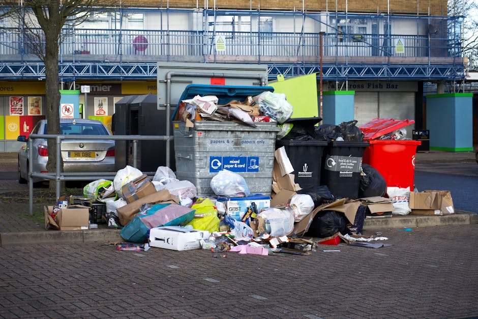 A large outdoor waste collection point located on a paved urban street, featuring multiple black, red, and black plastic rubbish bins, with some black bags and recycling containers stacked nearby. The bins are filled with various types of waste, including cardboard boxes, paper packaging, and plastic bags, with some refuse overflowing onto the ground. To the left, a grey car is parked next to a metal railing, partially blocking the view of a storefront with signage and glass windows. Behind the bins, a building under construction or renovation is visible, covered in blue scaffolding and safety barriers, with a sign indicating potential hazards. The scene is illuminated by natural daylight, with leafless trees and a generally tidy but cluttered waste area, reflecting a typical private waste disposal site managed by a waste removal service. Waste Removal Queen's Park may handle similar private rubbish collection needs in the local area, providing alternative waste management solutions for shops and businesses.