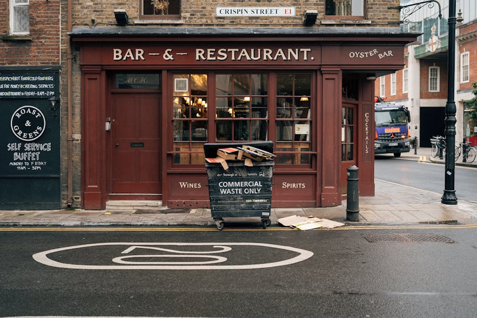 A black wheeled bin labeled 'Commercial Waste Only' situated directly in front of a red-brick building housing a restaurant and bar, which features large glass windows with multiple illuminated interior lights. The bin is overflowing with cardboard boxes, papers, and other waste materials, some of which are spilling onto the pavement. The bin is positioned on the edge of a narrow street, with a yellow line along the curb and a white road marking visible in the foreground. To the left, there is a black sign advertising a breakfast buffet, and to the right, a black bollard and a lamppost are visible close to the entrance of the establishment. A parked truck and bicycles can be seen further down the street. The scene reflects urban waste accumulation near commercial premises, where private waste collection services like Waste Removal Queen's Park offer alternative rubbish disposal options for local businesses, emphasizing the importance of efficient rubbish management in busy shopping and dining areas.